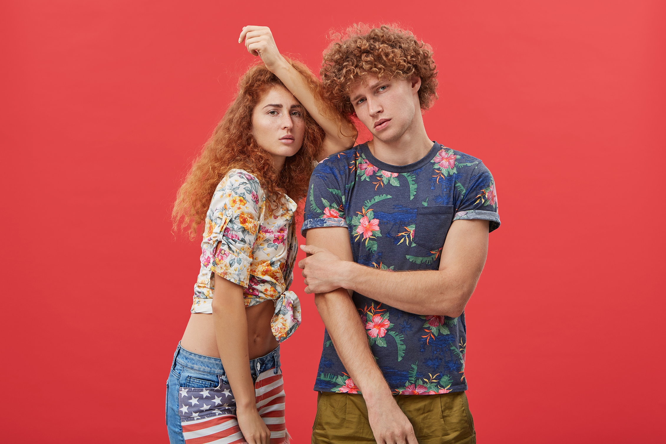 Beautiful woman with red hair and freckles wearing shirt and shorts leaning at shoulder of her curly boyfriend who is posing looking directly into camera with serious expression. Two best friends